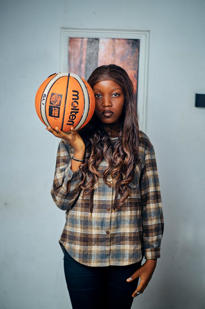 A confident young woman holds a basketball indoors, showcasing her athletic spirit and style.